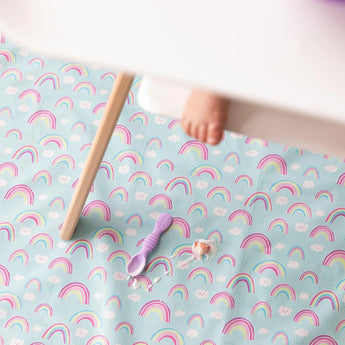 A baby's foot peeks from under a high chair tray, while Bumkins Splat Mat: Rainbows lays below with a purple spoon and yogurt smear on top.