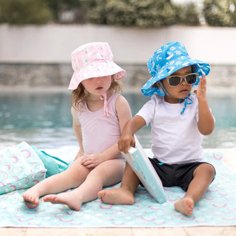 Two kids sit by a pool on Bumkins Splat Mat: Rainbows—one in a pink hat and swimsuit, the other in a blue hat, sunglasses, and white shirt.