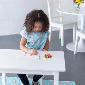 A curly-haired child in a rainbow smock draws at a white table with a Bumkins Splat Mat: Rainbow underneath.