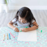 A girl draws on white paper with colored pens at a table covered by a Bumkins Splat Mat: Rainbows.
