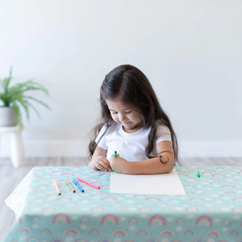 A young girl with long hair sits at a table covered by a Bumkins Splat Mat: Rainbows, coloring on paper with markers.
