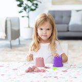 A girl plays with a purple sandcastle mold and kinetic sand on a Bumkins Splat Mat: Floral, with a plant and sofa in the background.