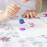 Close-up of a child painting with a blue sponge brush on a Bumkins Splat Mat: Floral.