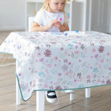 A child in a white shirt crafts at a table covered by a Bumkins Splat Mat: Floral in a bright room.