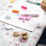 Close-up of a child drawing with markers on a Bumkins Splat Mat: Floral, with cookies nearby.
