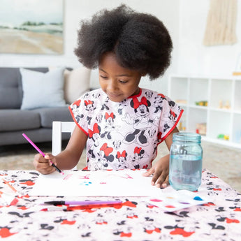 A child paints at a table covered by Bumkins Splat Mat: Minnie Mouse, wearing a matching smock, in a cozy living room.