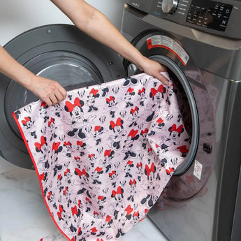 A person places Bumkins Splat Mat: Minnie Mouse into an open front-loading washing machine.