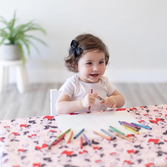 A toddler in a black bow draws with crayons at a table covered with Bumkins Splat Mat: Minnie Mouse.
