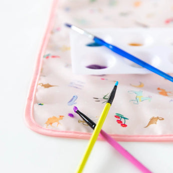 Close-up of three paintbrushes and a paint palette resting on Bumkins Splat Mat: Princess Magic on a white background.