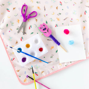 Craft supplies like scissors, paints, and brushes lie on a Bumkins Splat Mat: Princess Magic on a white background.