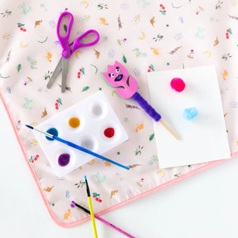 Craft supplies like scissors, paints, and brushes lie on a Bumkins Splat Mat: Princess Magic on a white background.