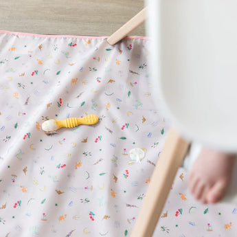 A yellow spoon with yogurt sits on top of Bumkins Splat Mat: Princess Magic underneath a high chair with a baby's foot visible.