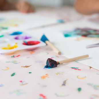 Close-up of colorful paint blobs, a blue-covered brush, and paper on a table covered by Bumkins Splat Mat: Princess Magic.
