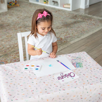 A girl with a pink bow paints with watercolors and brushes at a table with Bumkins Splat Mat: Princess Magic in a bright room.