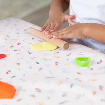 A child uses a wooden rolling pin to roll yellow clay on Bumkins Splat Mat: Princess Magic while cookie cutters sit close by.