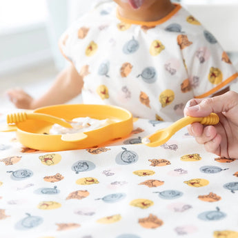 A child sits at table with a Bumkins Splat Mat: Winnie and Friends, with a dish of yogurt in front of them. An adult holds a spoon nearby.