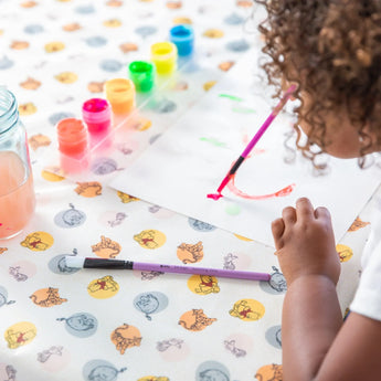 A child paints with watercolors at a table, with colorful paints and a water jar on Bumkins Splat Mat: Winnie and Friends for easy cleaning.