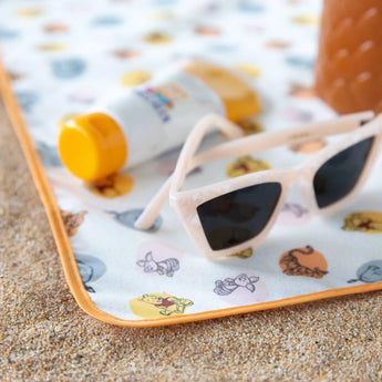 Close-up of sunglasses and sunscreen resting on a Bumkins Splat Mat: Winnie and Friends on sand.