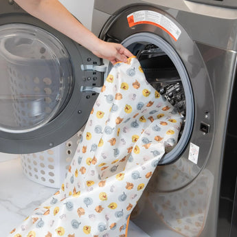 A person places Bumkins Splat Mat: Winnie and Friends in an open front-loading washing machine.