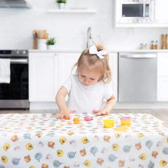 A toddler with a white bow plays with colorful clay on a Bumkins Splat Mat: Winnie and Friends in a bright kitchen.