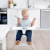 A baby sits in a high chair wearing a Bumkins Silicone Bib: Hearts, looking down at a gray bowl as they smile in a bright kitchen.