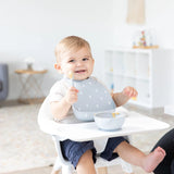 A happy baby in a high chair wears a Bumkins Silicone Bib: Hearts, holding a spoon by a gray food bowl in a bright room.