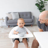 A baby in a high chair smiles with a spoon, wearing a Bumkins Silicone Bib: Hearts as an adult leans over; a gray sofa and plant are behind.