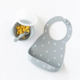 Bumkins Silicone Bib: Hearts lays next to a gray bowl & spoon set with scrambled eggs rests on a white background.