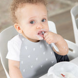 A curly-haired toddler with a messy face eats with a spoon, wearing a Bumkins Silicone Bib: Hearts.