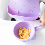 Close-up of a child in a Bumkins Silicone Bib: Rainbows enjoying mac and cheese from a matching purple bowl on a high chair tray.