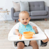 A baby in a high chair smiles during mealtime, wearing a Bumkins Silicone Bib: Ocean Life and eating from an orange bowl.