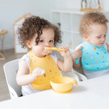 Two toddlers enjoy mealtime at a white table; one wears a Bumkins Silicone Bib: Camp Gear and eats from a yellow bowl with a spoon.