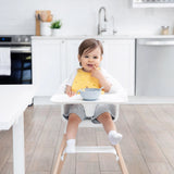 A baby in a high chair wears a Bumkins Silicone Bib: Camp Gear, smiling with a gray bowl on the tray in a modern kitchen.