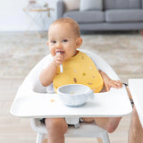 A baby in a high chair wearing a Bumkins Silicone Bib: Camp Gear eagerly tackles mealtime, enjoying food from a gray bowl with a spoon.