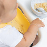 Close-up of a baby wearing a Bumkins Silicone Bib: Camp Gear, with mashed food in a bowl with a spoon nearby.