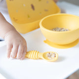 Close-up of a child wearing Bumkins Silicone Bib: Camp Gear, gripping a yellow spoon with food beside a matching bowl on a high chair tray.