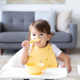 A toddler sits in a high chair wearing a Bumkins Silicone Bib: Camp Gear, enjoying baby-led weaning from a yellow bowl with a spoon.
