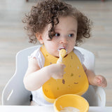 A curly-haired toddler with a messy face in a high chair wears a Bumkins Silicone Bib: Camp Gear, eating yogurt from a yellow bowl.