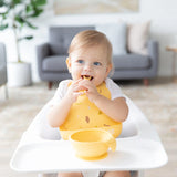 A smiling toddler sits in a high chair wearing a Bumkins Silicone Bib: Camp Gear, exploring self-feeding from a yellow bowl with a spoon.