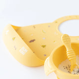 Close-up of Bumkins Silicone Bib: Camp Gear a yellow bowl with yogurt, and a yellow spoon on a white background.