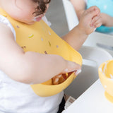 A toddler in a Bumkins Silicone Bib: Camp Gear messily enjoys creamy food as they reach into the front pocket of the bib.