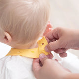 Hands fasten the closure of a Bumkins Silicone Bib: Camp Gear around a baby's neck.