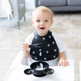 A smiling toddler in a high chair wears a Bumkins Silicone Bib: Mickey Mouse Faces, holding a spoon by a food-filled bowl on the tray.