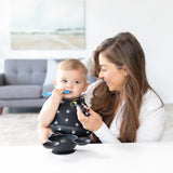 A woman smiles at a baby wearing a Bumkins Silicone Bib: Mickey Mouse Faces chewing a blue spoon in a bright, art-filled room.