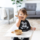 A toddler in a Bumkins Silicone Bib: Mickey + Minnie Mouse eats from a gold bowl in a high chair, with a gray couch behind.