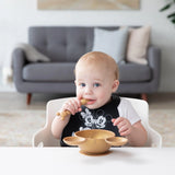 A baby in a high chair wears a Bumkins Silicone Bib: Mickey + Minnie Mouse, holding a spoon near a gold bowl; a gray sofa in the background.