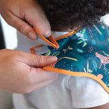 Adult hands fasten the hook-and-loop closure of Bumkins Super-Sized SuperBib®: Jungle around a child's neck.