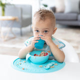 A toddler sips from a Bumkins Silicone Starter Cup: Blue at a white table, wearing a patterned bib with a matching bowl and food mat.
