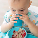 Close-up of a baby in a bib drinking from a Bumkins Silicone Starter Cup: Blue by a bowl of oatmeal.