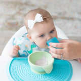 A baby in a bib and bow sits in a high chair learning to drink from a Bumkins Silicone Starter Cup: Blue, guided by an adult hand.